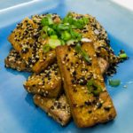 alt="a blue square plate with slices of sautéed sesame crusted tofu topped with chopped green onions."