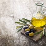 alt="a clear glass bottle with olive oil flanked by a couple of olive branches and a handfull marinated olives."