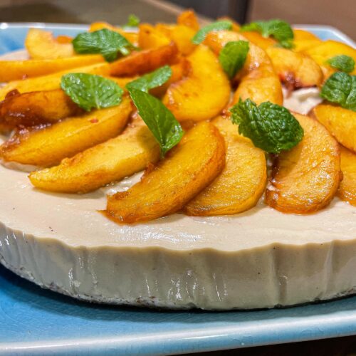 alt="a plant-based peach cheesecake decorated with fresh peaches and mint and served on a blue plate."