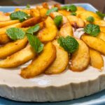 alt="a plant-based peach cheesecake decorated with fresh peaches and mint and served on a blue plate."