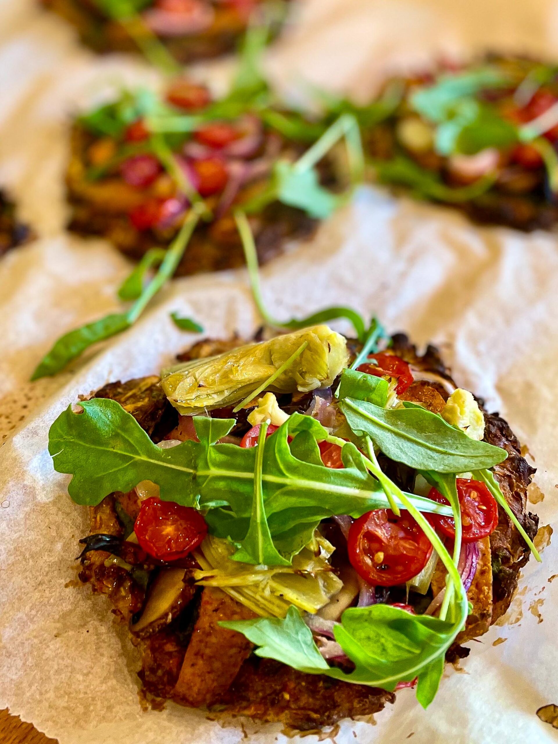 alt="a close-up of a mini root vegetable pizza topped with smoked tofu, cherry tomatoes, artochoke hearts and arugula."