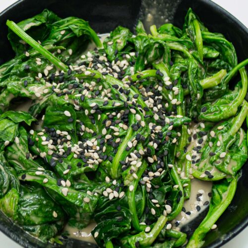 alt="a bowl with steamed spinach in a sesame sauce, sprinkled with black and white sesame seeds."