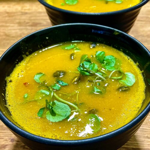 alt="a soothing looking yellow soup topped with green watercress and served in a black bowl."