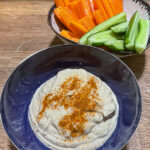 alt="a bowl of hummus served with sticks of carrot and cucumber."