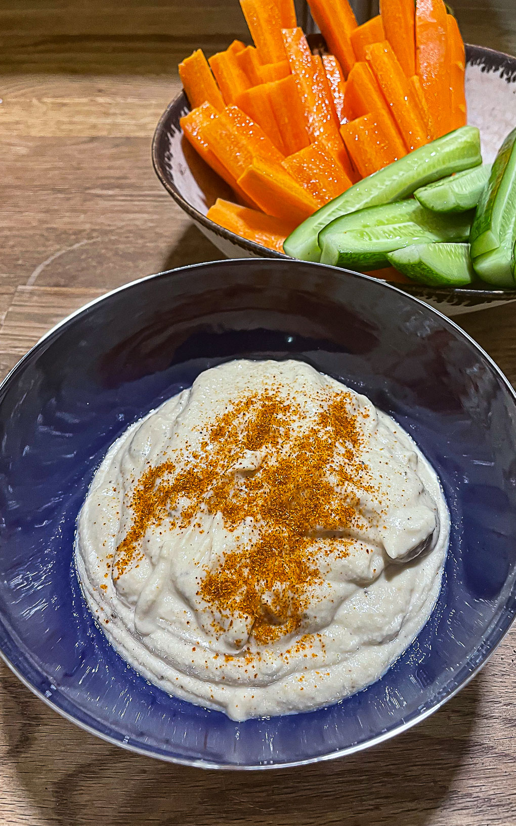 alt="a bowl of hummus sprinkled with cayenne pepper and served with sticks of carrot and cucumber."