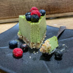 alt="a slice of green matcha mousse cake on a black plate. It is topped with raspberries and blueberries. A bite of cake on a fork sits next to it on the plate."