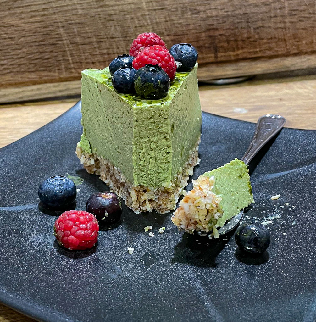 A slice of green matcha mousse cake on a black plate. It is topped with raspberries and blueberries and a bite of cake on a fork sits next to it on the plate.
