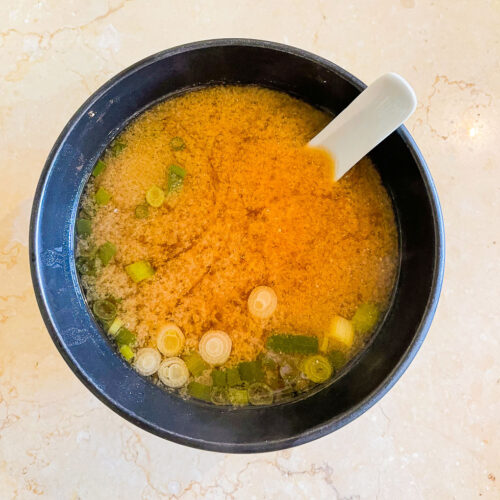 alt="a bowl of miso soup with green onions and a ceramic spoon."
