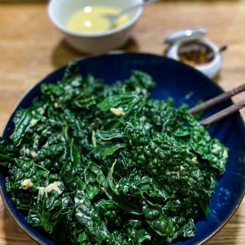 alt="deeply green Tuscan kale drizzled with zesty dressing and served in a dark blue bowl. Extra dressing and chili flakes on the side."