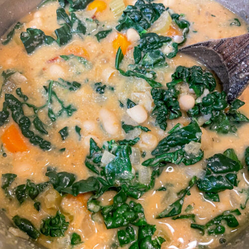 alt="a pot of white beans and kale soup with a wooden stir spoon in it."