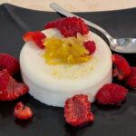alt="a white lemongrass coconut panna cotta with lemon relish and raspberries served on a black plate."
