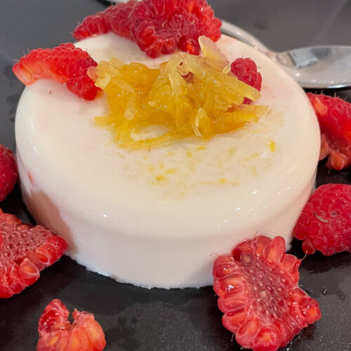 alt="a white lemongrass coconut panna cotta with lemon relish and raspberries served on a black plate."
