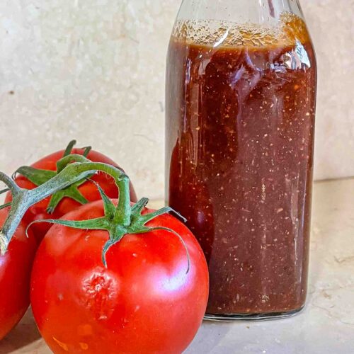 alt="a bottle of homemade ketchup sat next to three plump tomatoes."